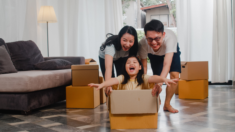 happy asian young family having fun laughing moving into new home japanese parents mother father smiling helping excited little girl riding sitting cardboard box new property relocation